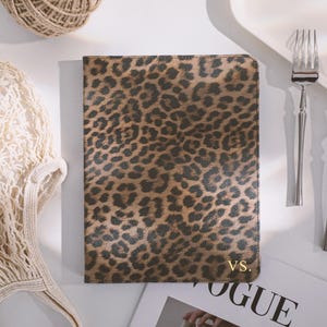 Può includere: Un diario con stampa leopardata e le iniziali dorate "VS." è mostrato su una superficie bianca. Una forchetta e un coltello sono posti a destra del diario. Una rivista Vogue, un gomitolo di spago e una borsa a rete sono anche nell'immagine.