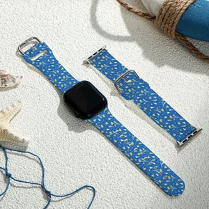 May include: Blue watch band with a repeating pattern of yellow and white floral designs. The watch band is shown with a black watch face and a silver buckle. A second band is also shown. The background includes seashells and a life preserver.