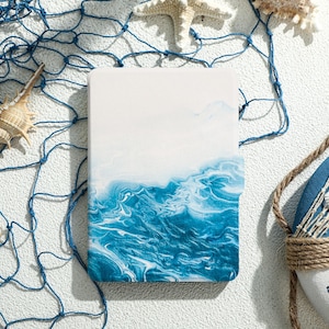 Könnte beinhalten: Ein weißes und blaues Notizbuch mit einem Aquarelldesign von Ozeanwellen. Das Notizbuch ist umgeben von einem blauen Fischernetz, Muscheln und einem Rettungsring.