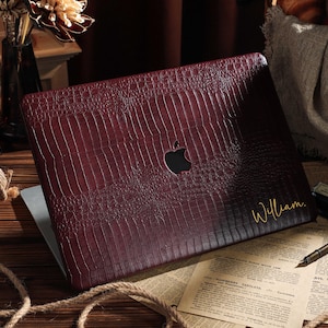 Pode incluir: Uma capa de laptop bordô com um padrão texturizado semelhante ao de um crocodilo. A capa apresenta um recorte para o logotipo da Apple e o nome "William" em escrita dourada. O laptop está aberto, revelando uma tela prateada.