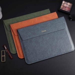 Peut inclure: Trois pochettes pour ordinateur portable en cuir vert, orange et bleu. La pochette bleue porte le nom "Diana" en relief doré. Les pochettes sont disposées sur une surface noire, avec un verre d'eau, du maquillage et des lunettes de soleil en arrière-plan.