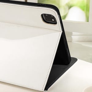 Può includere: Una custodia in pelle bianca con una patta nera per un tablet o un telefono. La custodia è piegata per mostrare l'interno nero.