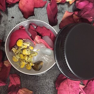 Könnte beinhalten: Ein kleiner, runder, silberner Behälter gefüllt mit getrockneten Rosenblättern, Kamillenblüten und einem klaren Kristall. Der Behälter ist von getrockneten Rosenblättern auf einem grauen Hintergrund umgeben.