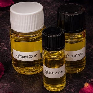 Könnte beinhalten: Drei klare Glasflaschen gefüllt mit gelber Flüssigkeit, beschriftet mit 'Glücksel' und den Volumina 20 ml, 10 ml und 5 ml. Die Flaschen haben weiße und schwarze Schraubverschlüsse und sind von dunklen rosa Rosenblättern und einem Kristall umgeben.
