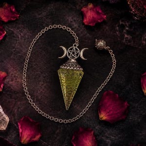 Könnte beinhalten: Ein spitzes, facettiertes lindgrünes Kristallpendel, das an einer silberfarbenen Metallkette hängt. Die Spitze des Kristalls ist mit einer dekorativen silbernen Kappe versehen, die ein zentrales Pentagramm-Symbol zwischen zwei nach außen gerichteten Mondsicheln zeigt.