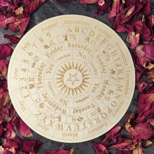 Könnte beinhalten: Ein rundes Wahrsagebrett aus Holz mit eingraviertem Text und Symbolen, einschließlich Monate, Tage und Alphabet. Das Brett hat ein zentrales Sonnendesign und ist von getrockneten Rosenblättern umgeben. Das Brett hat einen Durchmesser von etwa 25 cm.