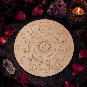 Könnte beinhalten: Eine runde, hellfarbene Holzplanchette mit eingravierten Buchstaben, Zahlen und Symbolen. Die Planchette hat ein zentrales Sonnenstrahl-Design und ist von Text umgeben, darunter Monate, Tage und das Alphabet. Die Planchette wird zur Wahrsagung verwendet.