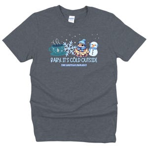May include: Dark gray t-shirt with a winter-themed graphic. The design includes a steaming teal mug, a snowflake, a snake wearing a hat and scarf, and a snowman. Text reads "Baby, It's Cold Outside" and "The Reptile Project."