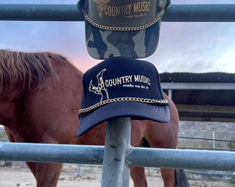 Może przedstawiać: Dwie czarne i zielone czapki trucker z złotymi łańcuchami i napisem "COUNTRY MUSIC made me do it" z przodu. Czapki wiszą na metalowym słupku ogrodzenia.