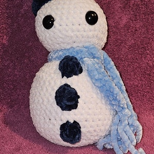 May include: A white crocheted snowman with a blue scarf and a black velvet top hat. The snowman has three black buttons and black eyes.