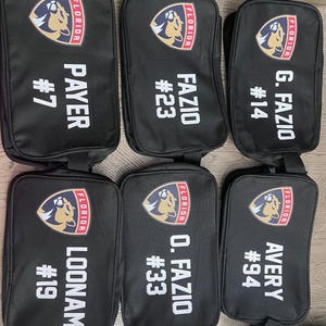 Peut inclure: Six sacs à dos noirs à fermeture éclair avec un logo des Panthers de la Floride et des noms et numéros de joueurs. Les sacs sont alignés en rangée sur une table.