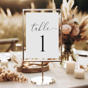 Puede incluir: Una tarjeta de número de mesa blanca con un marco dorado y un soporte plateado. La tarjeta dice "mesa 1" en letra cursiva negra.