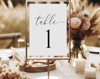 May include: A white table number card with a gold frame and a silver stand. The card says "table 1" in black cursive font.