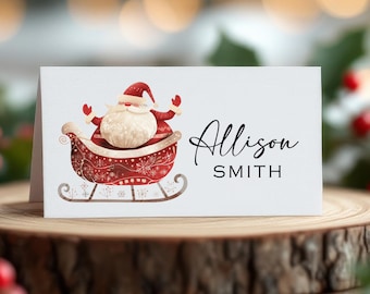 May include: A white folded place card featuring a watercolor illustration of Santa Claus in a red sleigh with snowflake details. The name "Allison SMITH" is printed in black script and block letters. The card is displayed on a wooden surface.
