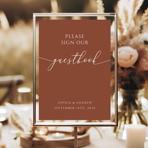 May include: A sign with a brown background and white text that reads "Please sign our guestbook" with the date "September 14th, 2025" and the names "Sophia & Andrew" below. The sign is in a silver frame and is sitting on a table.