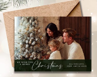 May include: Christmas card featuring a family decorating a Christmas tree. The card has a green banner with the words "We wish you a Merry Christmas" and "All our love, John, Amy & Louis". The card is inside a brown envelope.