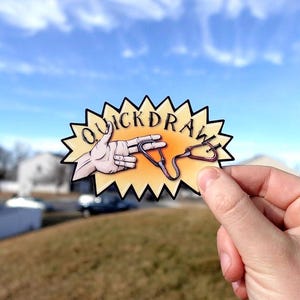 May include: A sticker with a jagged yellow border featuring the word "QUICKDRAW" in black letters. The sticker depicts a hand holding a climbing quickdraw. The background shows a blue sky and a grassy hill.