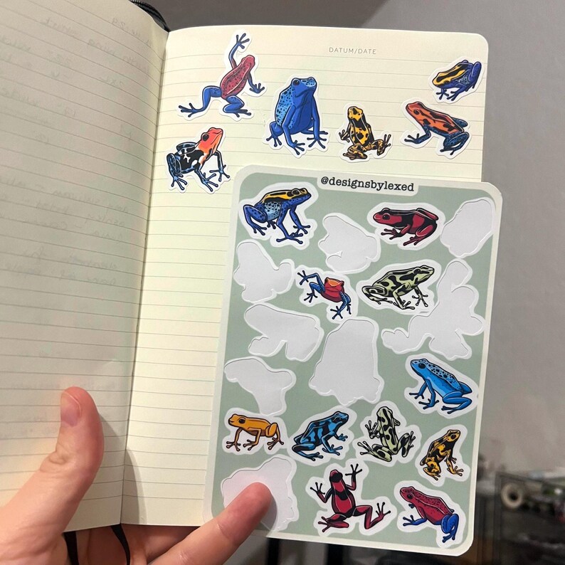 May include: A sheet of colourful frog stickers with a variety of colours and patterns. The stickers are arranged in a grid pattern on a white background. The text "@designsbylexed" is printed in black at the top of the sheet.