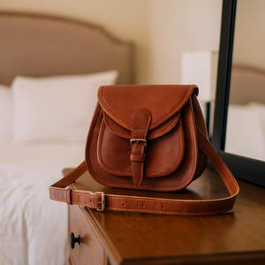 Könnte beinhalten: Eine braune Ledersatteltasche mit Klappenverschluss und verstellbarem Schulterriemen. Die Tasche hat eine Schnalle und wird auf einer Holzoberfläche präsentiert. Die Tasche ist etwa 25 cm breit.