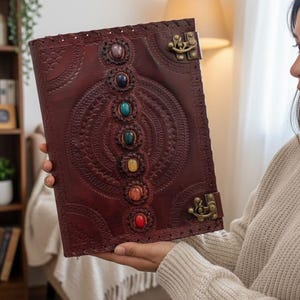 May include: A dark brown leather journal with embossed patterns and a row of colorful gemstones. The journal has a decorative clasp and is held open, showcasing the intricate design and craftsmanship. A unique, handcrafted item.