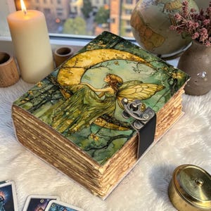 May include: A handmade journal with a fairy illustration on the cover. The cover art features a fairy sitting on a crescent moon, with a green and gold color scheme. The journal has a metal clasp and a leather strap.