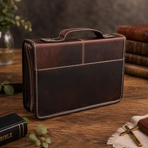 May include: A rectangular dark brown leather case with a top handle and a wrap-around zipper closure. The case features visible light-colored stitching along the edges and seams, with a smooth, matte finish and a structured, boxy design.