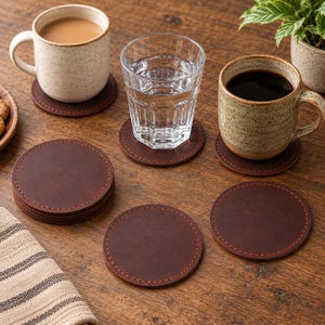 Peut inclure: Un ensemble de dessous de verre circulaires en cuir marron foncé avec des coutures orange visibles sur les bords. Les dessous de verre sont disposés sur une surface, certains placés individuellement et d'autres empilés, servant de support.
