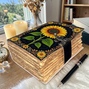 May include: A handmade journal with a sunflower design on the cover. The cover is black with a large yellow sunflower and green leaves. The pages are aged, and the journal is secured with a black strap and metal clasp. A black pen rests nearby.