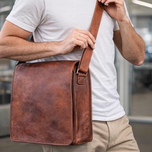 以下が含まれることがあります： フラップ開閉と調節可能なショルダーストラップが付いた、茶色の革製メッセンジャーバッグ。 バッグは長方形で、ヴィンテージ感を出すために、使い古したような仕上がりになっています。 ストラップは金属製の金具で取り付けられています。