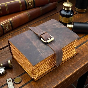 Puede incluir: Un diario rectangular con cubierta de cuero marrón oscuro y una correa de cuero a juego asegurada por una hebilla de metal. El diario contiene páginas gruesas, texturizadas y de color tostado claro con bordes irregulares visibles.