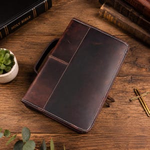 May include: A dark brown leather zippered cover featuring a side handle and white contrast stitching. The rectangular case has a smooth, distressed finish with visible grain patterns and a structured design, providing a protective exterior for books.