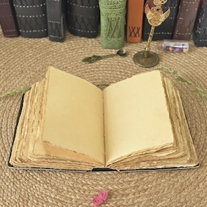 Puede incluir: Un diario abierto de estilo antiguo con p&aacute;ginas envejecidas de color crema. El libro tiene una cubierta negra y una encuadernaci&oacute;n visible. Las p&aacute;ginas tienen un borde rugoso y rasgado. El diario est&aacute; sobre una superficie tejida, de color beige, con otros objetos decorativos en el fondo.