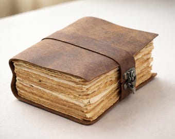 May include: A brown leather journal with a strap closure and metal clasp. The pages are aged, giving the appearance of an antique book. The journal is rectangular and appears to be handmade.