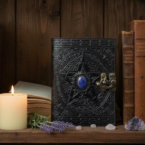 Puede incluir: Diario de cuero negro con un dise&ntilde;o de pentagrama y una gema azul en el centro. El diario tiene un cierre ornamentado. Una vela encendida, libros y cristales lo rodean, creando una est&eacute;tica m&iacute;stica.