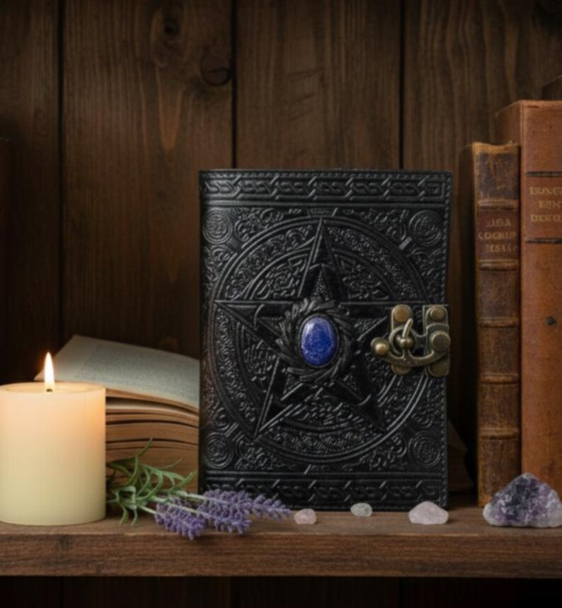 Puede incluir: Diario de cuero negro con un dise&ntilde;o de pentagrama y una gema azul en el centro. El diario tiene un cierre ornamentado. Una vela encendida, libros y cristales lo rodean, creando una est&eacute;tica m&iacute;stica.