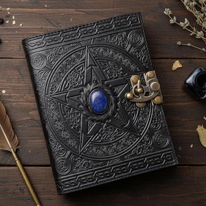 Puede incluir: Diario de cuero negro con dise&ntilde;o de pentagrama en relieve y una gema azul en el centro. El diario presenta un cierre de metal ornamentado y detalles de nudos celtas. Un accesorio de escritura decorativo.