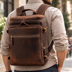 May include: A dark brown leather backpack featuring a rectangular silhouette, multiple zippered compartments, and brass-toned hardware. The bag includes a top handle, adjustable shoulder straps, and side D-ring attachments with visible stitching.