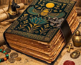 May include: A large, antique-style book with a teal cover featuring gold and teal Egyptian-inspired designs. The book has aged, yellowed pages and a black leather strap closure. Additional Egyptian artifacts are in the background.