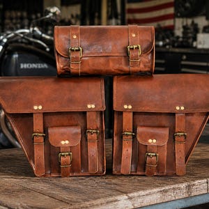 May include: Three brown leather motorcycle bags with brass buckles and straps. The bags are stacked on a wooden surface in front of a motorcycle. The bags have a vintage, rustic appearance.