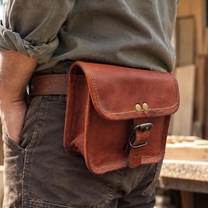 May include: A brown leather belt pouch featuring a fold-over flap with two brass rivets and a metal buckle closure. The rectangular bag is attached to a matching brown leather belt worn around the waist, showcasing visible stitching along the edges.