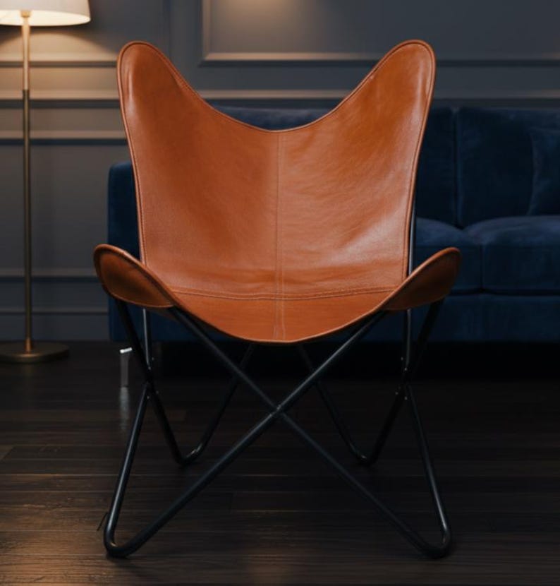 Op de afbeelding: Een bruine leren vlinderstoel met een zwart metalen frame. De stoel heeft een kenmerkende vleugelvormige rugleuning en zitting en staat tegen een donkere achtergrond. De stoel is een stijlvol meubelstuk.