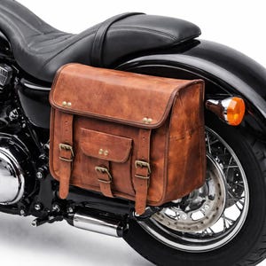 Può includere: Una borsa da sella per moto in pelle marrone con fibbie e rivetti in ottone è montata sul lato di una moto nera. La borsa ha una forma rettangolare con chiusura a patta e una piccola tasca frontale.