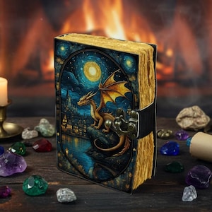 May include: A fantasy journal with a dragon illustration. The cover depicts a golden dragon overlooking a city at night, under a full moon. The journal has a metal clasp and aged pages. The pages appear to be approximately 20 cm in height.