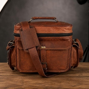 Könnte beinhalten: Eine braune Kameratasche aus Leder mit einem Tragegriff, einem schwarzen Reißverschluss und einer Vordertasche mit Klappe, die mit zwei messingfarbenen Nieten und einem Schnallenriemen gesichert ist. Die Tasche hat Seitentaschen und einen verstellbaren Schultergurt.