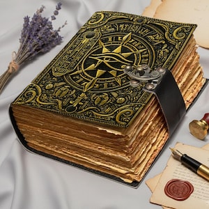Op de afbeelding: Een gedetailleerd, antiek ogend dagboek met een zwart en goudkleurig omslag met een oogsymbool en kompasroos ontwerp. Het dagboek is vastgezet met een zwarte leren riem en sluiting. De verouderde pagina's suggereren een vintage esthetiek.