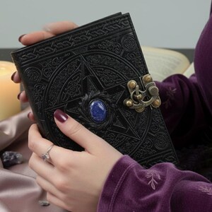 Puede incluir: Diario de cuero negro con dise&ntilde;o de pentagrama en relieve, una gema azul y un cierre decorativo. El diario est&aacute; en las manos de una persona, con un anillo en un dedo. El fondo incluye una vela encendida y un libro.