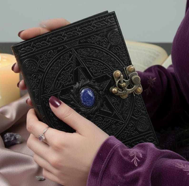 Puede incluir: Diario de cuero negro con dise&ntilde;o de pentagrama en relieve, una gema azul y un cierre decorativo. El diario est&aacute; en las manos de una persona, con un anillo en un dedo. El fondo incluye una vela encendida y un libro.