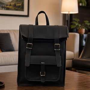 May include: A black rectangular backpack featuring a fold-over top flap, a top carry handle, and two vertical straps with metal buckles. A smaller front pocket is secured with a central vertical strap and a metal buckle, all in a matte finish.