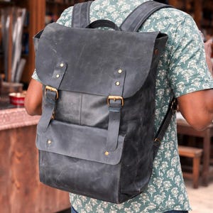 May include: A black leather backpack with a flap closure and adjustable straps. The backpack features brass-colored buckles and rivets. The bag is worn over the shoulders and has a distressed finish, suggesting a vintage or rugged style.