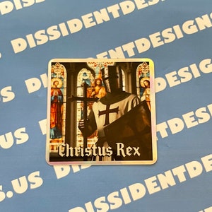 Può includere: Un adesivo argentato con finitura olografica che presenta un cavaliere in armatura che tiene una spada e una croce. Il cavaliere è in piedi davanti a una finestra di vetro colorato. Il testo "Christus Rex" è stampato sull'adesivo.